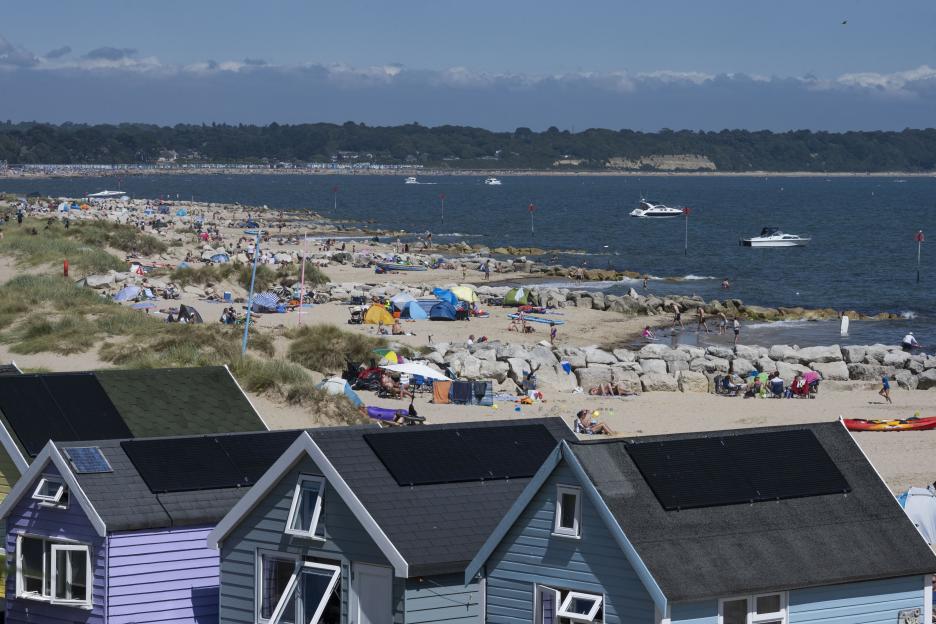 Beach huts with solar panels overlook a crowded beach at Mudeford, Dorset.