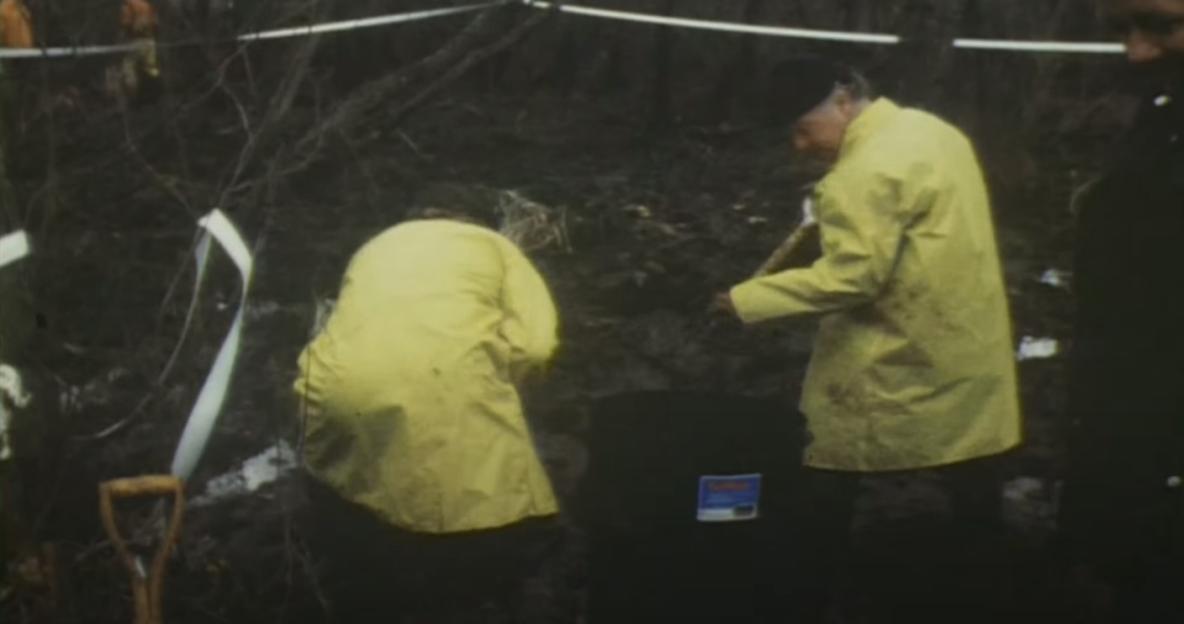 Two people in yellow raincoats digging in a muddy, wooded area with a white tape boundary.