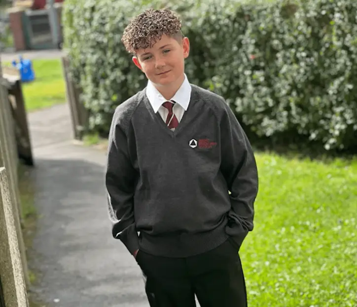A young boy in a school uniform stands with his hands in his pockets.