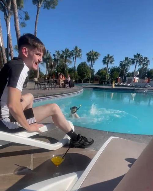 A man in a black and white shirt sits on a lounge chair by a pool while a person in snorkeling gear splashes in the water.