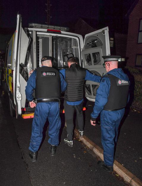 An arrested man in a black vest is escorted into a police van by two Greater Manchester Police officers.