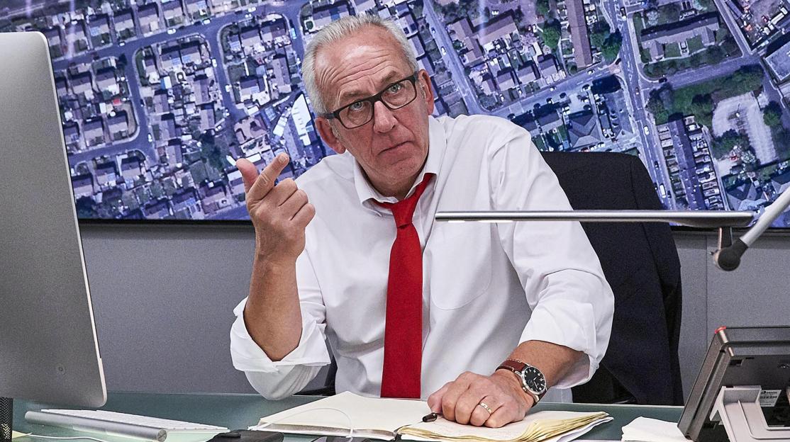 Peter Bleksley wearing glasses and a red tie, gesturing towards a map of a town.