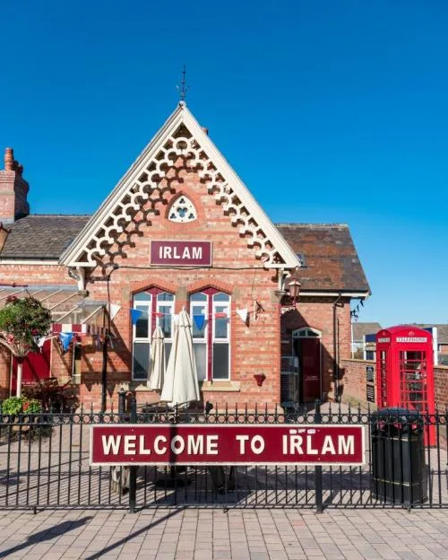 Exterior view of the Irlam station building, featuring a "WELCOME TO IRLAM" sign and a red telephone box against a clear blue sky.