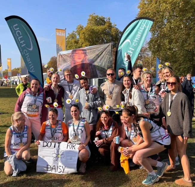 A group of runners wearing medals and daisy headbands pose for a photo behind a banner reading "Do It For Daisy".