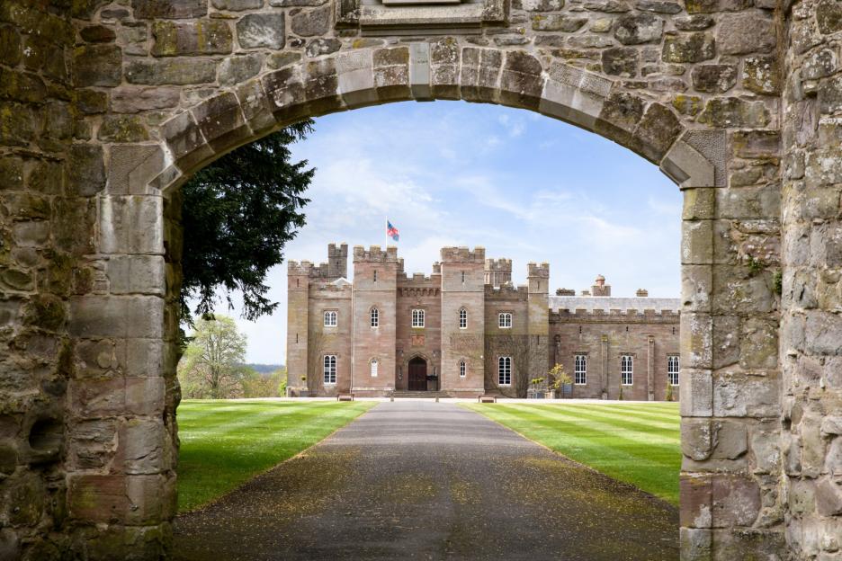 A stone archway frames a long driveway leading to a large historic castle with multiple towers and a flag flying from one.