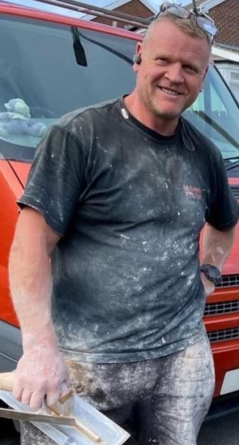 Nick, a construction worker, stands smiling in front of a red van, holding a trowel.