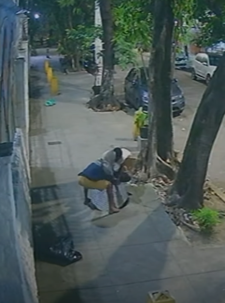 A person bends down on a sidewalk, next to a black garbage bag.
