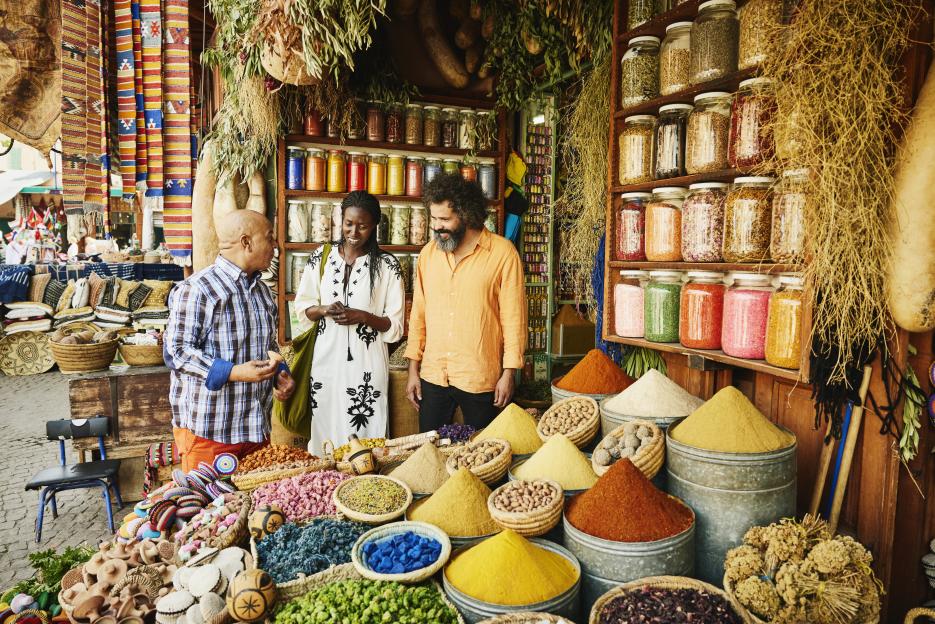 A couple talks to a vendor in a spice shop in the souks of Marrakech, with colorful spices in jars and piles.