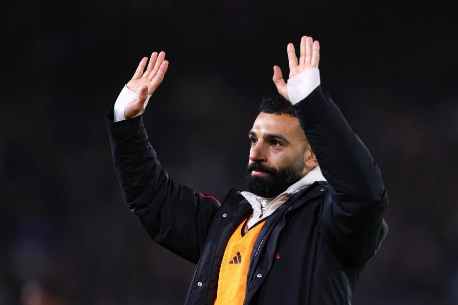 Salah raising his hands, wearing a black jacket over an orange Adidas bib.