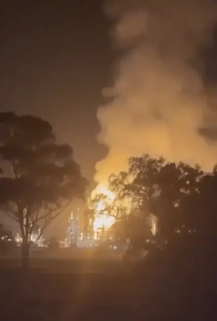A large fire and smoke plume rising from an industrial facility at night, silhouetting trees in the foreground.