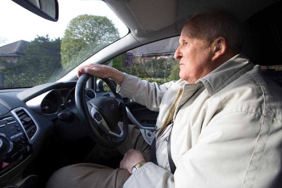 An elderly man with hearing aids sits in the driver's seat of a car, holding the steering wheel.