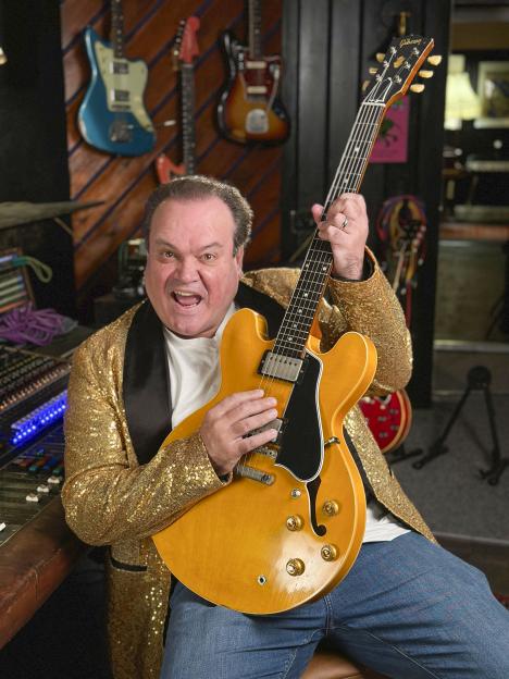 Shaun Williamson performing in a gold sequined jacket, holding a Gibson electric guitar and singing.