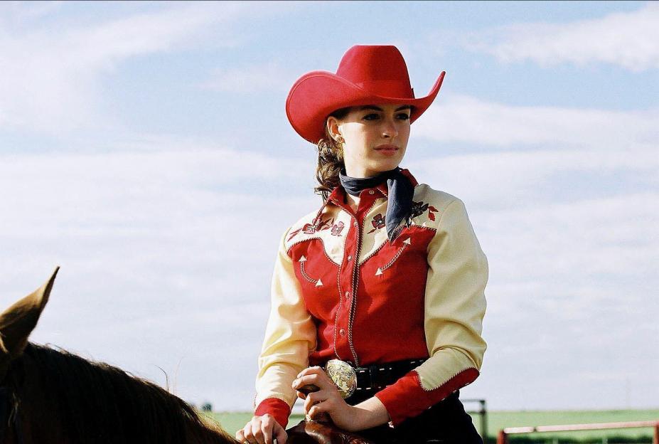 Anne Hathaway as Lureen Newsome in Brokeback Mountain, wearing a red cowboy hat, red and cream shirt, and a black scarf while on horseback.