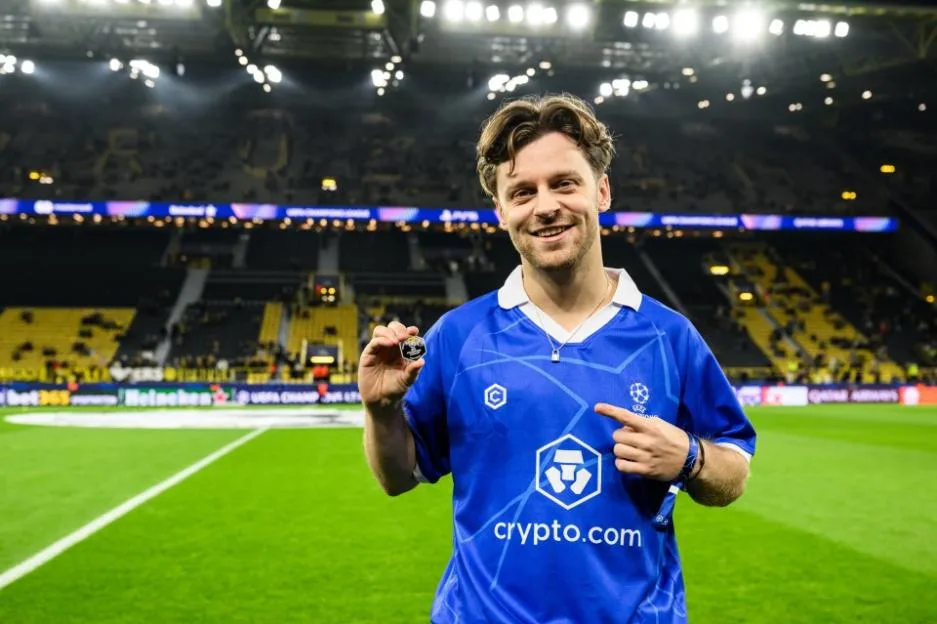 A man in a blue jersey with a Crypto.com logo holds up a commemorative coin on a football pitch.