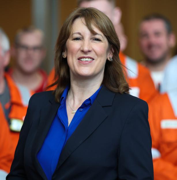 Britain's Chancellor Rachel Reeves smiling in a blue shirt and black blazer in front of workers in orange jumpsuits.