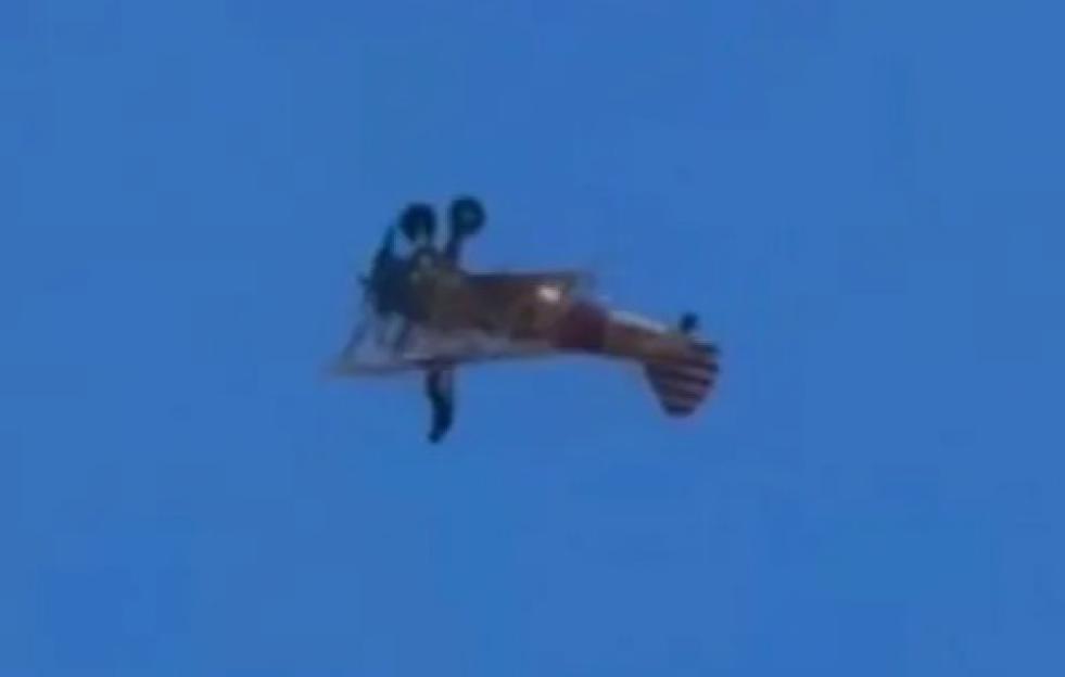 A biplane flies through a clear blue sky, with a white cloud visible in the upper left corner.