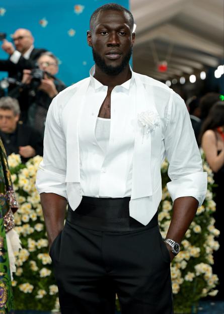 Stormzy at the Met Gala wearing an open white shirt, white vest, black trousers, and a white boutonniere.