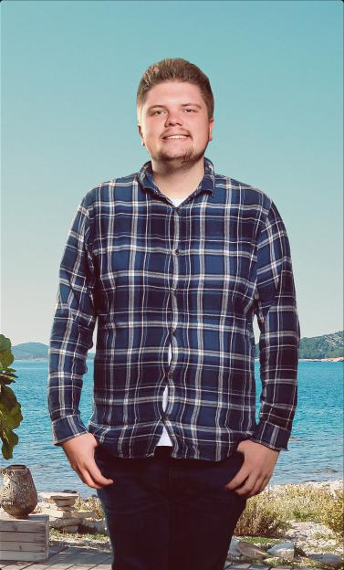 A man wearing a blue plaid shirt stands smiling against a backdrop of the ocean and hills.
