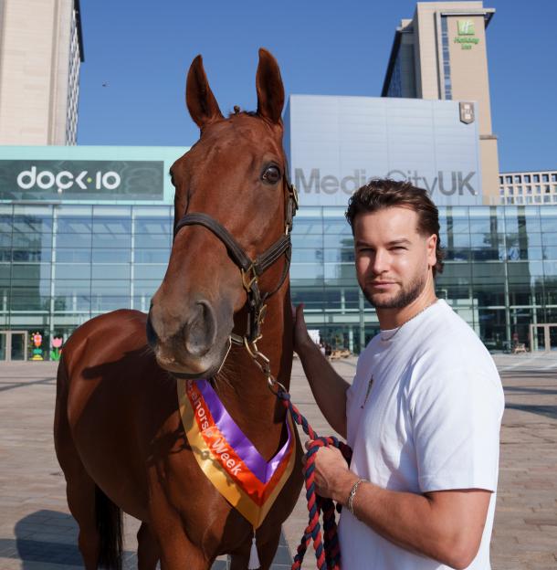 A brown racehorse with a "Racehorse Week" ribbon and a man in a white shirt stand in front of modern buildings.