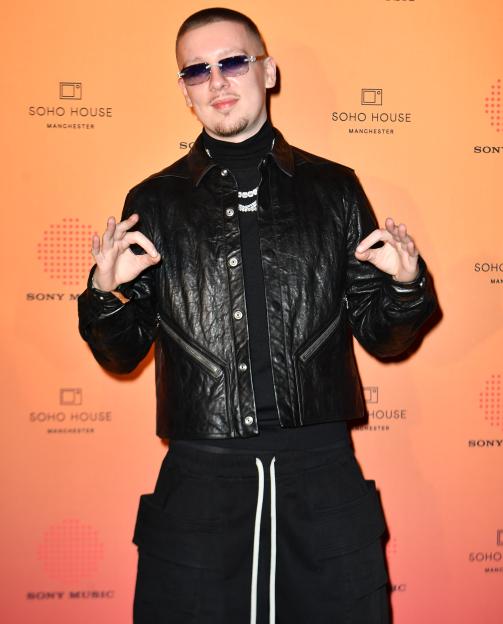 Rapper Aitch wearing a black leather jacket, turtleneck, and dark cargo pants, making an "OK" gesture with both hands at the Brit Awards afterparty.