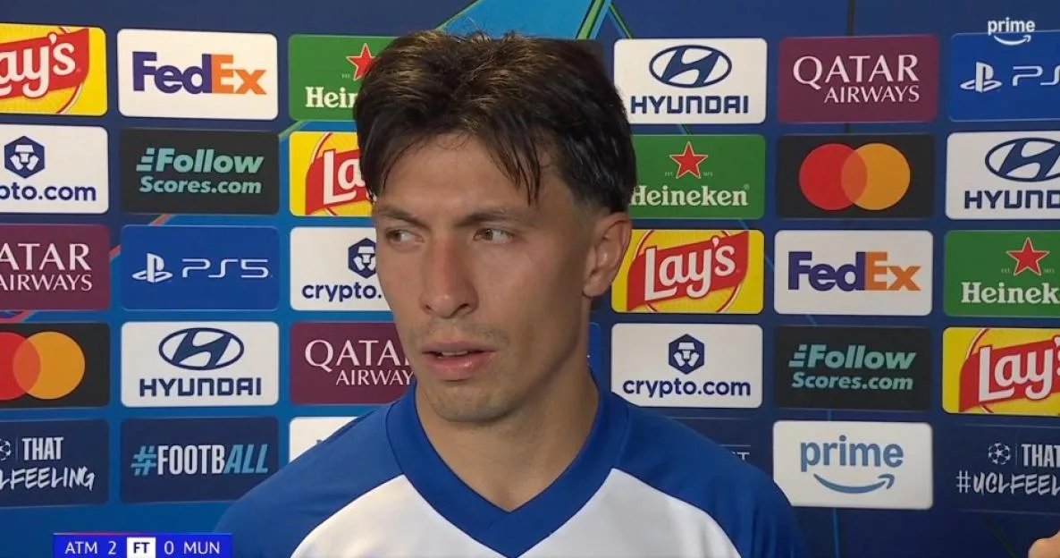 A footballer in a blue and white kit is interviewed in front of a sponsor backdrop.