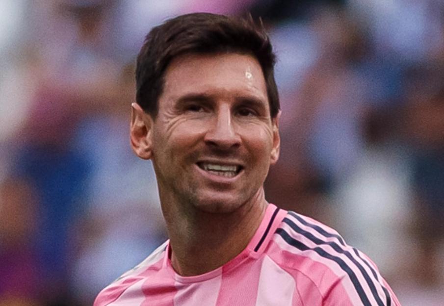 Lionel Messi in a pink and white striped Inter Miami jersey smiles on the field.