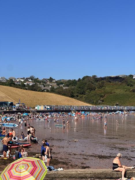 A crowded beach with many people walking in shallow water and sitting on the sand, with a long line of colorful beach houses behind them, and houses on a hill in the distance.
