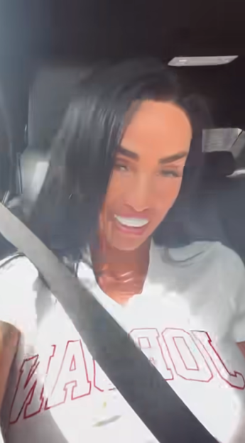 A smiling woman with dark hair and a white t-shirt with red lettering, wearing a seatbelt in a car.