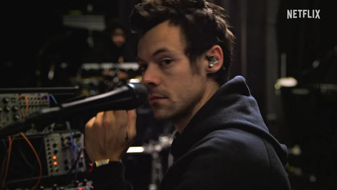 A man with an in-ear monitor holds a microphone and looks over his shoulder.