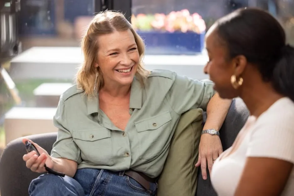 Joanna Page smiling and holding a device while sitting on a couch and talking with another woman.
