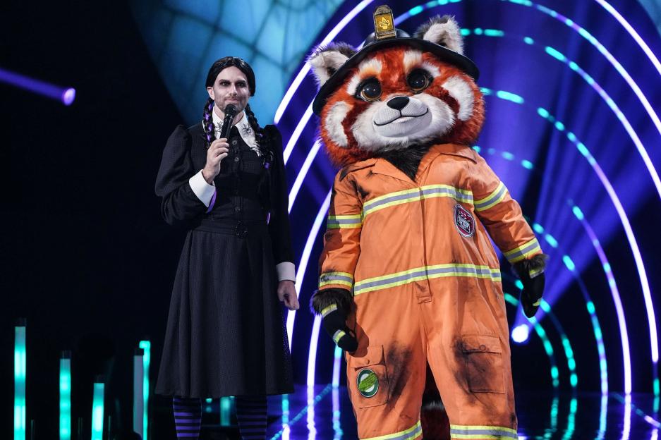 Joel Dommett dressed as Wednesday Addams standing next to a person in a Red Panda firefighter costume on The Masked Singer.