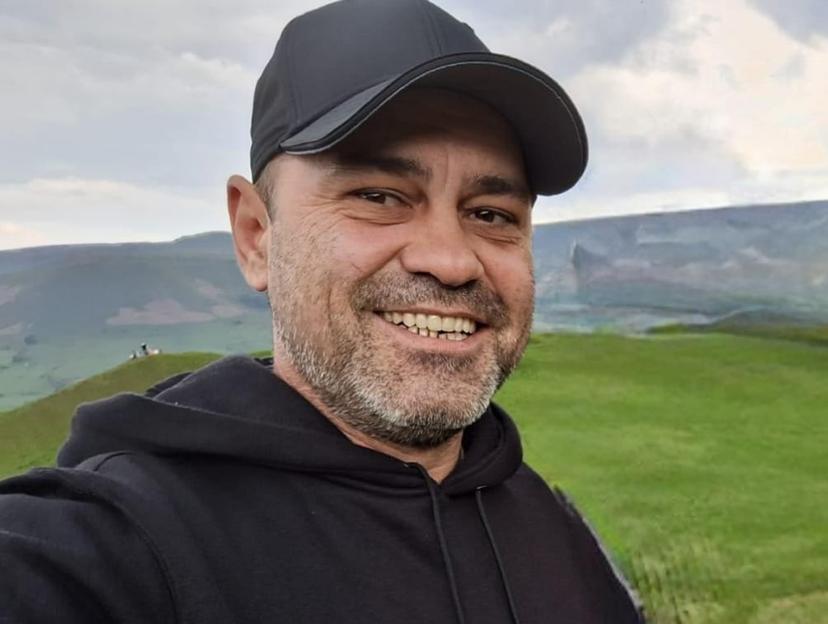 Portrait of Claudiu-Carol Kondor smiling, wearing a black cap and hoodie, with a green hillside and cloudy sky in the background.