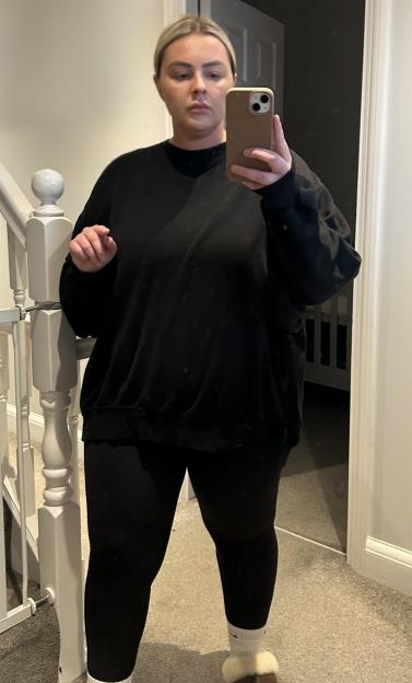 Amy Salmon before her weight loss, standing in a mirror wearing a dark top, leggings, and slippers.