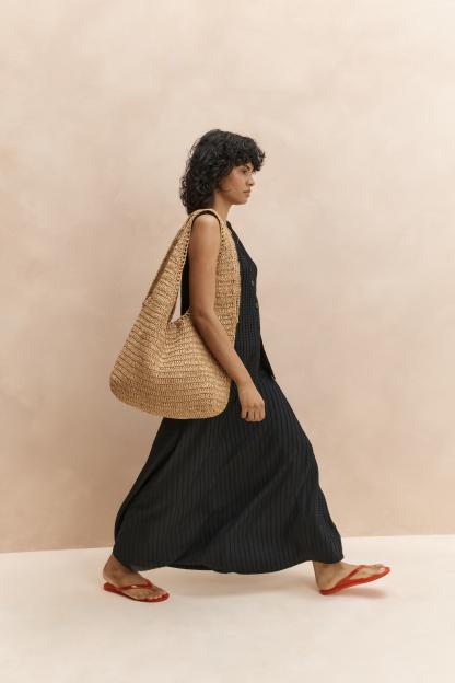 A woman in a black maxi dress and red flip-flops carries a woven straw tote bag.