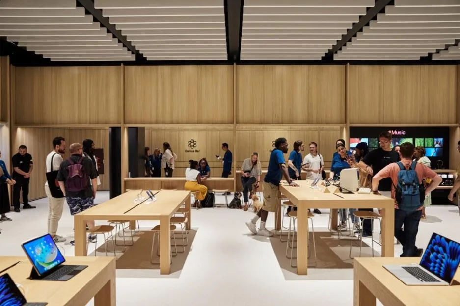 A spacious Apple store with light wooden walls and tables, with multiple customers and employees interacting.
