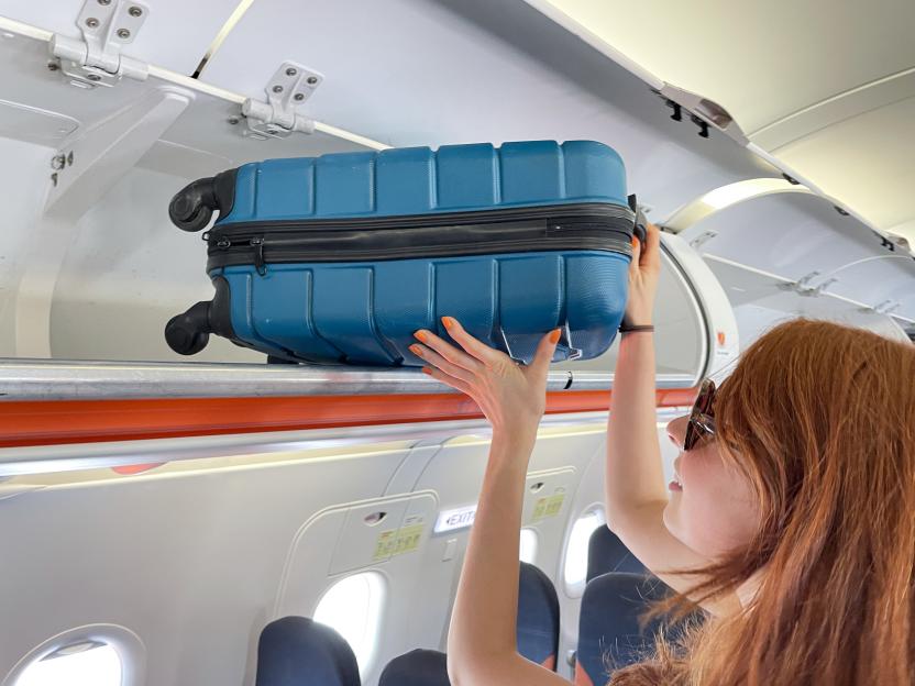 Close-up image of attractive, redheaded woman wearing sunglasses, placing wheelie suitcase in airplane cabin overhead baggage compartment, row of seats, focus on foreground