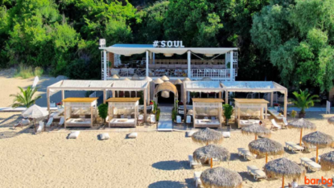 A beach club with cabanas and straw umbrellas on a sandy beach, backed by lush green trees, with a building named #SOUL in the background.