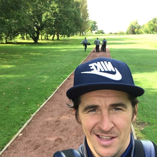Joey Barton wearing a baseball cap and smiling, with golfers in the background.