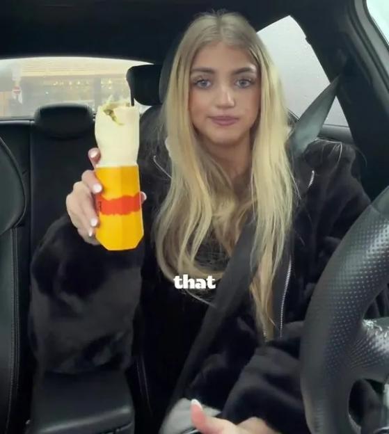 A woman with blonde hair in a car holds up a McDonald's wrap.