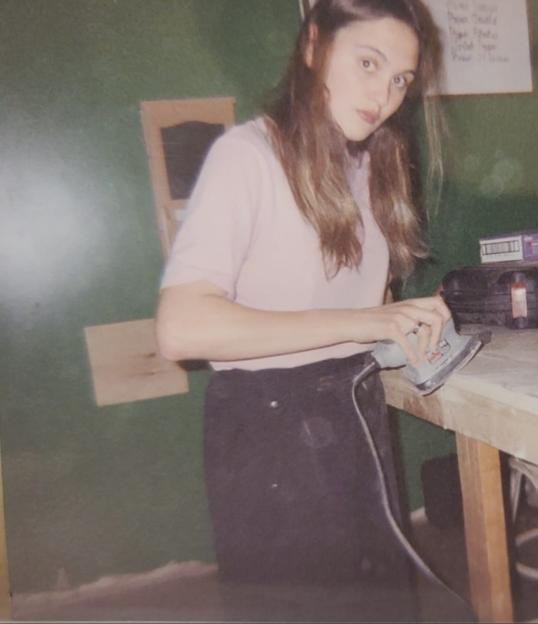 Two young women in pink shirts working at a workbench, one using a sander.