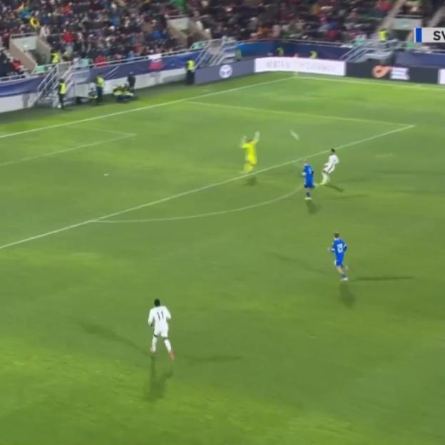 A soccer player in white dribbles past a player in blue as a goalie in yellow defends the goal.