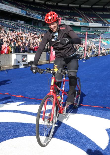 Greg James crossing the finish line on a red bicycle at Murrayfield Stadium for the Radio 1 Longest Ride challenge.