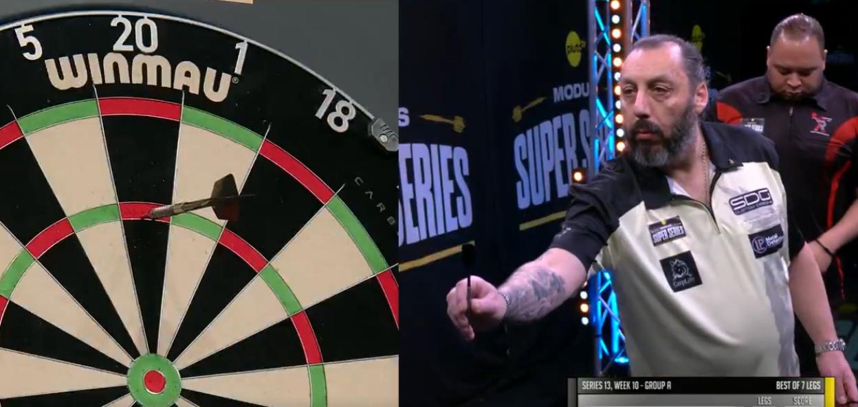 A dartboard with a dart stuck in the red portion of the number 1, and a darts player holding a dart on the right side of the image, with another man behind him.
