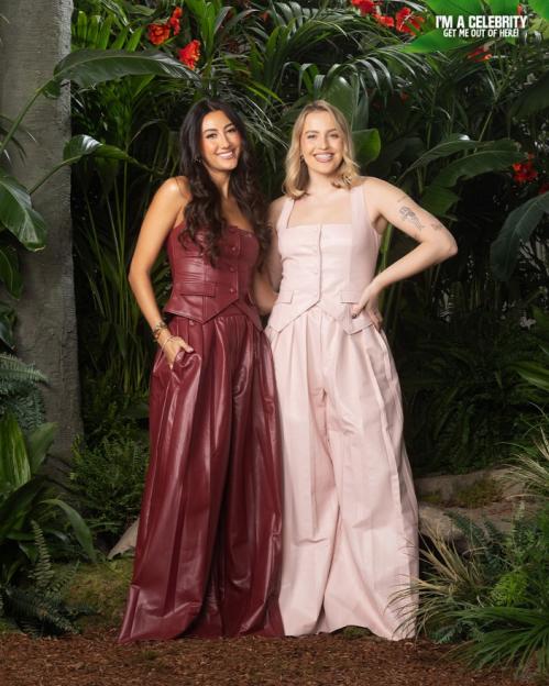 Two women in wide-legged pantsuits, one in burgundy and one in pink, posing in a jungle setting.