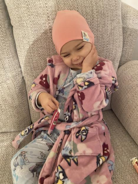 Primrose Hocking, a young child, wearing a pink beanie and a pink robe, smiling while holding a small packet of candy.