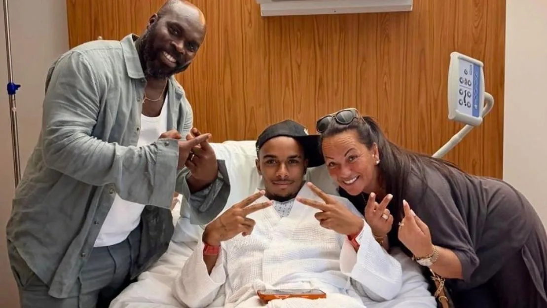 Three people smiling while posing together in a hospital room, with the person in the middle sitting in a hospital bed.