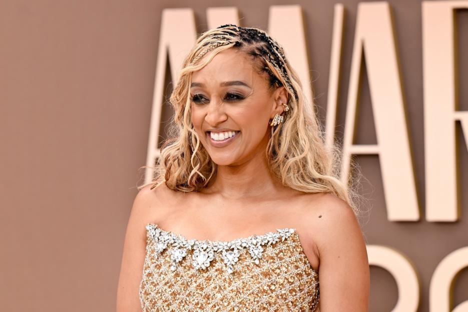 Tia Mowry smiles while wearing a gold, strapless, sequined gown with a jeweled neckline.