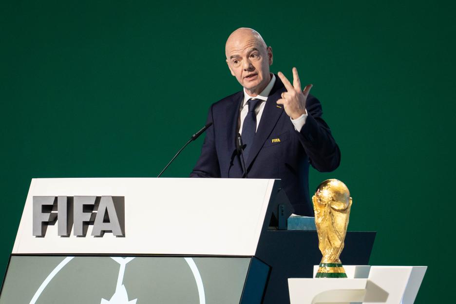 FIFA President Gianni Infantino speaking at a podium with the World Cup trophy nearby.