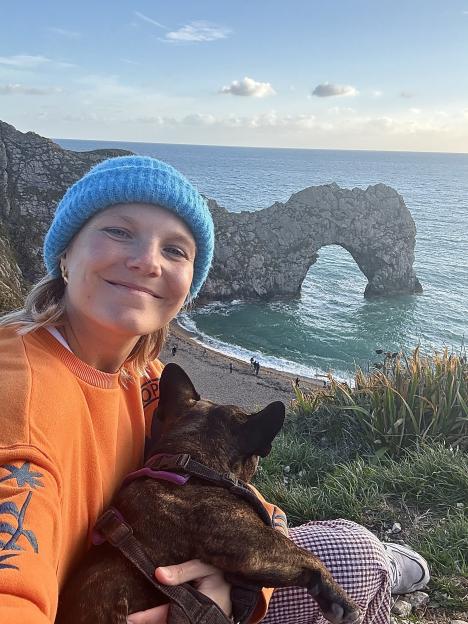 A woman in an orange sweater and blue beanie holds a brindle dog, smiling at the camera with the Durdle Door rock formation and ocean in the background.
