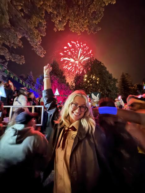 A woman in glasses and a dark jacket with a red tie cheers with a raised fist as red fireworks explode behind her at night.